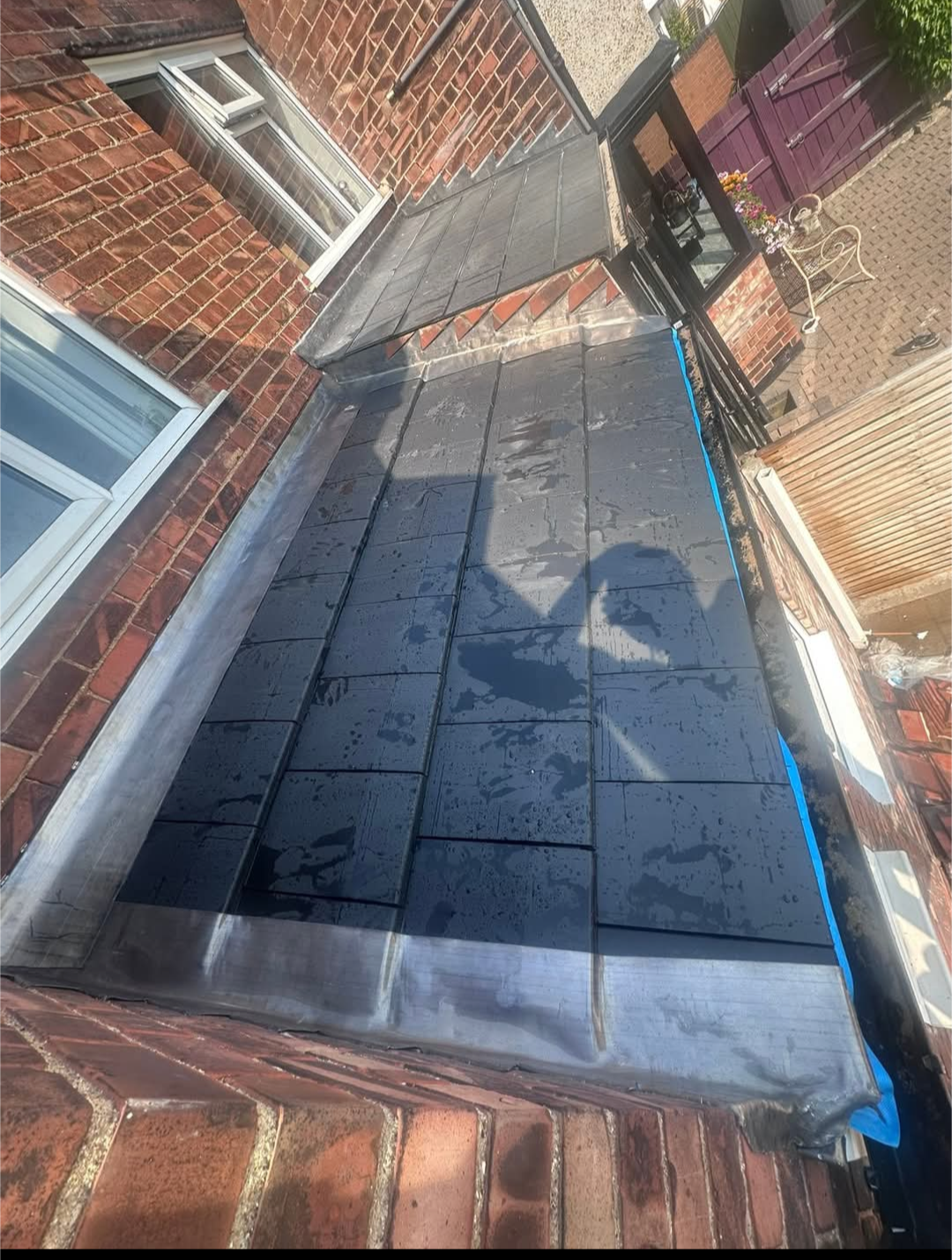 Completed bay roof with dark slates and lead flashings against a brick wall.
